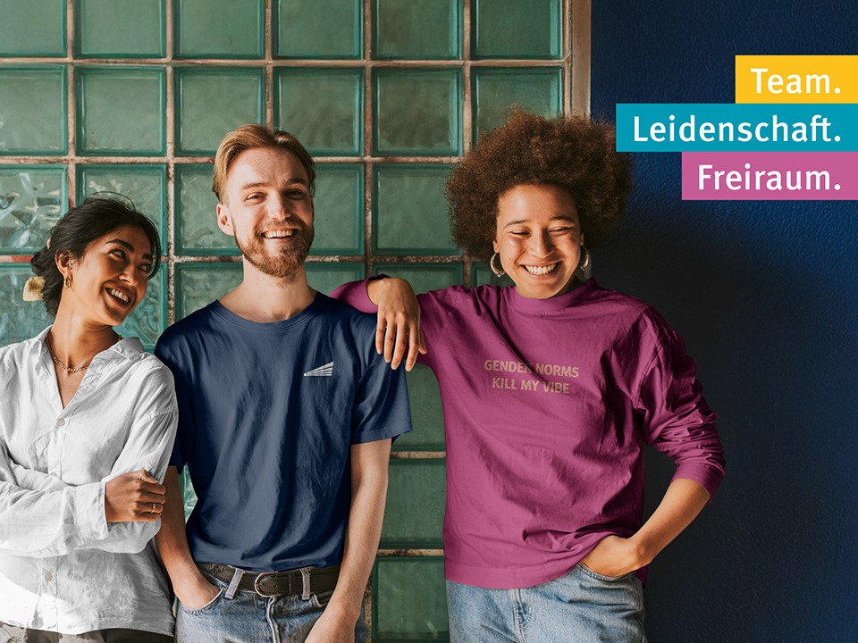 Das Bild wirbt für: "Pädagogische Fachkräfte – Karriere auf der Didacta 2026 Köln" Auf dem Bild stehen drei Personen nebeneinander und lachen gemeinsam. Links trägt eine Frau ein weißes T-Shirt. In der Mitte steht ein junger Mann mit blonden Haaren. Rechts ist eine Frau mit Afrofrisur zu sehen.